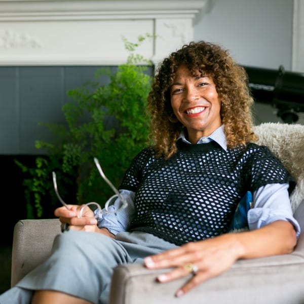 Julie Tinker sits on a chair holding her glasses and smiles 