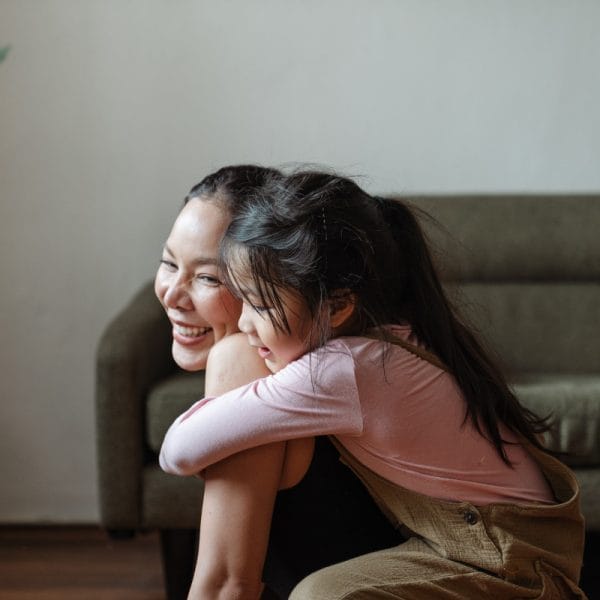 Picture of a young girl hugging her mother while sitting on a couch. The mother smiles.